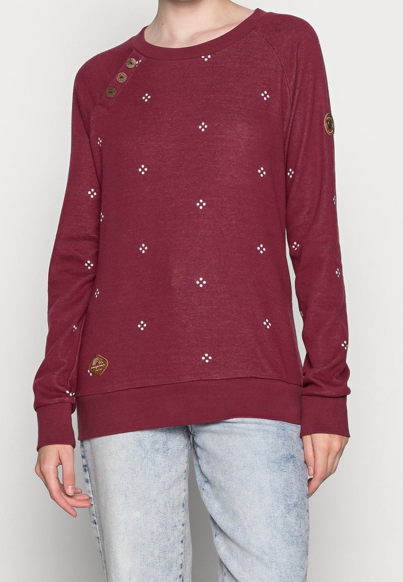 Personne portant un haut à manches longues bordeaux avec de petits motifs à pois blancs, des boutons sur l'épaule et un jean bleu clair.