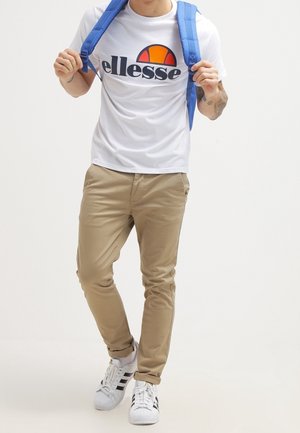 Junge Person trägt ein weißes ellesse-T-Shirt, beige Hose, weiße Adidas-Sneaker und einen blauen Rucksack, hält die Träger mit beiden Händen fest.