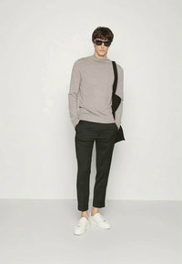 Man wearing gray long-sleeve top, black cropped pants, white sneakers, black shoulder bag, and sunglasses standing against white background.