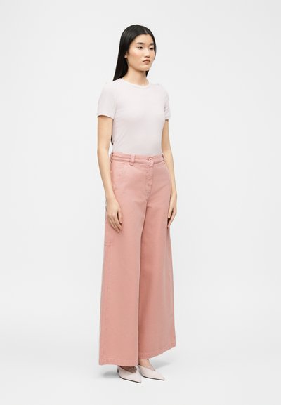 Γυναίκα όρθια, φοράει λευκό πουκάμισο με κοντά μανίκια, ροζ παντελόνι ψηλόμεσο με φαρδιά μπατζάκια και ανοιχτόχρωμα μυτερά παπούτσια, μπροστά σε απλό φόντο.