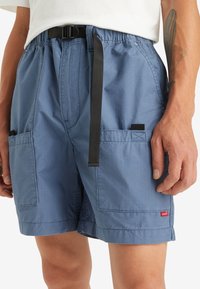 Shorts bleus en tissu léger, avec une taille élastique, une ceinture ajustable, et deux poches latérales avec un petit logo de marque.