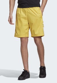 Gula sportshorts med elastiskt midjeband, sidfickor och vitt tre-stripes mönster på sidorna. Bärs med svarta sneakers.