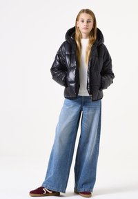 Zwarte pufferjack met een glanzende textuur, capuchon en ritssluiting, gecombineerd met losse blauwe denim jeans en bordeauxrode sneakers met witte accenten.