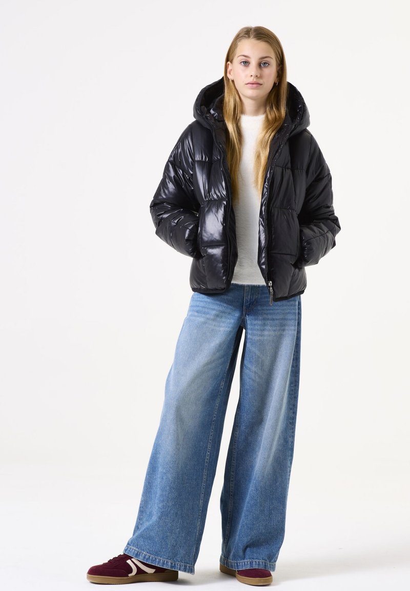 Zwarte pufferjack met een glanzende textuur, capuchon en ritssluiting, gecombineerd met losse blauwe denim jeans en bordeauxrode sneakers met witte accenten.