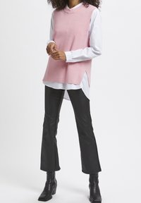 Maglione senza maniche rosa chiaro a maglia con un collo alto, indossato sopra una camicia bianca a maniche lunghe. Abbinato a pantaloni neri a zampa d'elefante e stivali neri.