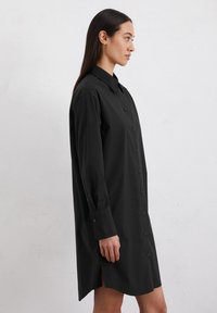 Zwarte knoopjurk met lange mouwen, een puntige kraag, zijslitsen en een gladde stofstructuur. Heeft een volledige knoopsluiting aan de voorkant.