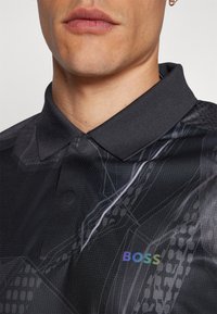 Melna polo krekls ar teksturētu virsmu, kuram ir abstrakti pelēki raksti un daudzkrāsains 'BOSS' logo kreisajā pusē.