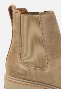 Bocanci Chelsea din suede de culoare cărămizie, cu panouri laterale elastice texturate, având o talpă din cauciuc și detalii de cusătură contrastante. Branding-ul Burkenstock este vizibil.