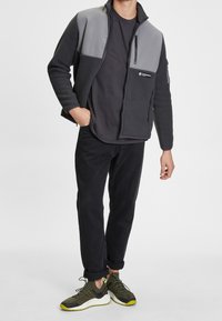 Zwarte fleecejas met grijze panelen, ritssluitingen en een hoge kraag. Gecombineerd met zwarte broek en olijfgroene sneakers met gele accenten.