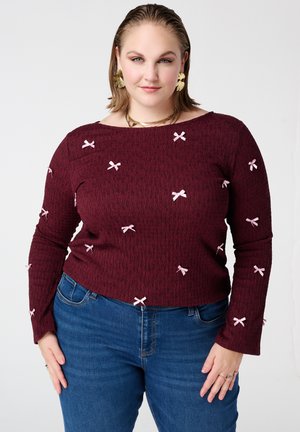 Vrouw draagt een kastanjebruine top met lange mouwen, kleine roze strikjes en een blauwe spijkerbroek, gecombineerd met gouden oorbellen en een ketting, staand met handen bij de heupen.