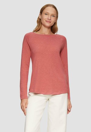 Femme avec des cheveux rouges lisses portant une chemise rouge délavée à manches longues et un pantalon blanc, se tenant devant un fond clair. 
