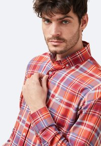 Camisa de cuadros rojos y azules con cuello abotonado, que presenta una tela texturizada y detalles de botones en contraste.