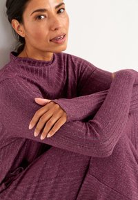 Langärmliges, geripptes, lila Kleid mit hohem Kragen und Daumenlöchern, das einen lockeren Schnitt und strukturierte Stoffqualität aufweist.