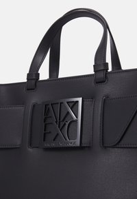 Black faux leather tote bag featuring a textured surface, double handles, and a prominent black logo panel on the front.