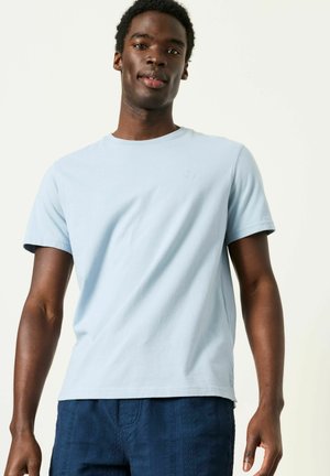 Mladý muž oblečený v světle modrém tričku s krátkými rukávy a tmavě modrých texturovaných kalhotách, stojící před jednobarevným světlým pozadím.