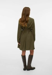 Robe chemise vert olive avec une ceinture à nouer, manches longues et ourlet arrondi. Associée à des bottes en cuir marron foncé à talons hauts.