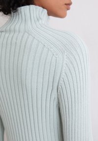 Frau in einem hellblauen, gerippten Rollkragenpullover, von hinten und zur Seite vor einem schlichten hellen Hintergrund gezeigt.