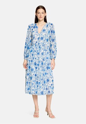 Kobieta stoi, ubrana w białą midi sukienkę z długim rękawem, z niebieskim kwiatowym wzorem oraz beżowe buty na otwartym obcasie na jednolitym tle.