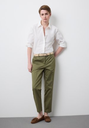 Persona con cabello corto, que lleva una camisa blanca de manga larga, pantalones verde oliva, cinturón beige y mocasines marrones, de pie contra una pared blanca.