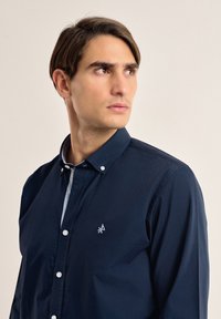Camisa azul oscuro de botones con corte ajustado, que presenta un cuello en contraste de color azul claro y un pequeño logotipo en el lado izquierdo del pecho. Material de algodón.