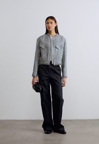 Γκρίζο κοντό μπουφάν με δύο μπροστινές τσέπες, ασημένιο φερμουάρ και ξεφτισμένες άκρες, σε συνδυασμό με μαύρο παντελόνι με φαρδιά πόδια και μια μικρή μαύρη τσάντα.