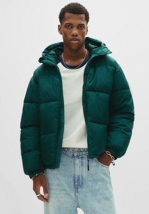 Grüne Pufferjacke mit Kapuze, ausgestattet mit einem gesteppten Design und elastischen Bündchen. getragen über einem hellen Sweatshirt und blauen Jeans.