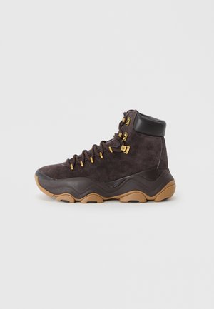 Brown suede high-top boot with black leather accents and gold eyelets, featuring a textured rubber sole and rounded toe design.