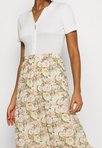 Valkoinen lyhythihainen toppi, jossa ribbineulos ja napit yksityiskohtina, yhdistetty pehmeissä pastellisävyissä olevaan kukkakuviolliseen hameeseen, jossa vihreitä lehtiä.