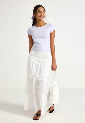 Młoda kobieta w jasnofioletowej koszulce, białej warstwowej maxi spódnicy, czarnych sandałach, trzymająca białą torebkę z falbankami, uśmiechająca się, stojąca na białym tle.