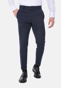 Pantaloni tailored blu navy realizzati in un tessuto leggero e liscio. Presentano tasche laterali, orli a fondo stretto e si posizionano appena sopra mocassini neri.