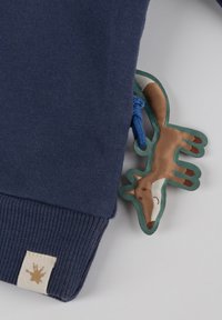 Manica blu navy con polsino a costine, etichetta beige con logo marrone e un portachiavi in stoffa a forma di volpe attaccato da un anello blu.