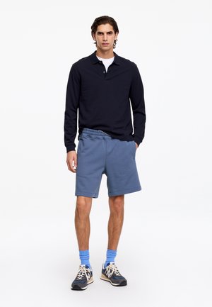 Mand stående iført en sort langærmet polo, blå shorts, blå sokker og sorte og hvide sneakers på en hvid baggrund.