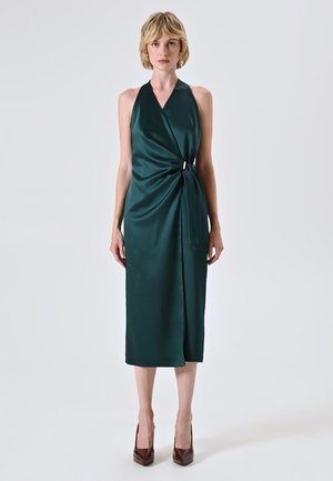 Satinkleid mit Halteroberteil in tiefem Türkis mit Wickeldesign, das an der Taille mit einem goldenen Akzent gebunden ist und bis zur Mitte der Wade fällt. Dazu passen High-Heels.