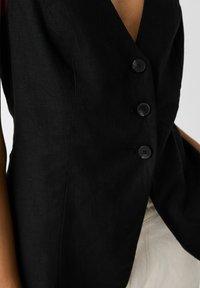 Svart linväst med V-ringning, som har tre svarta knappar och en skräddarsydd passform. Slät textur och minimal detaljering.