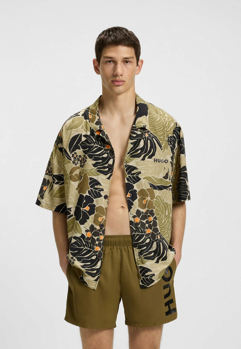 Korte mouw knoopshirt met een veelkleurig bloemenpatroon op een beige achtergrond, gecombineerd met olijfgroene shorts met een logo.