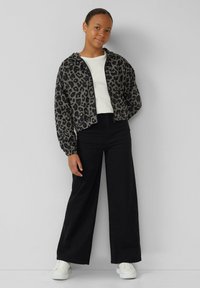 Geknipte jas met luipaardprint in grijs en zwart, gecombineerd met een witte blouse en zwarte wijde broek; model draagt witte sneakers.