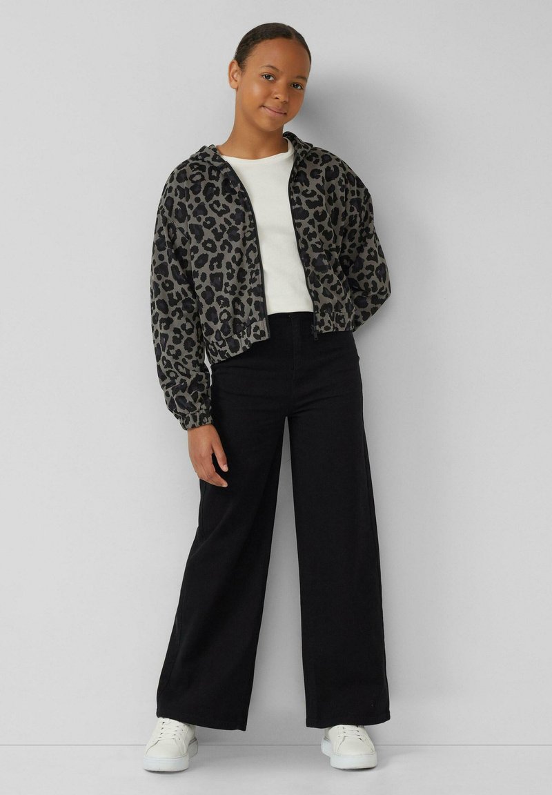 Geknipte jas met luipaardprint in grijs en zwart, gecombineerd met een witte blouse en zwarte wijde broek; model draagt witte sneakers.
