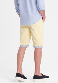 Pantaloni corti in cotone giallo chiaro con orlo risvoltato, abbinati a una camicia azzurro chiaro con bottoni e scarpe slip-on nere con suole bianche.