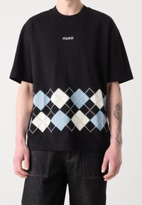 Schwarzes Baumwoll-T-Shirt mit einem weißen "HUGO"-Logo und einem Diamantmuster in Blau, Weiß und Schwarz am unteren Rand. Lockere Passform, kurze Ärmel.