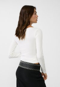 White long-sleeve top with a square neckline, paired with a black skirt featuring a studded belt accent. Smooth fabric texture.