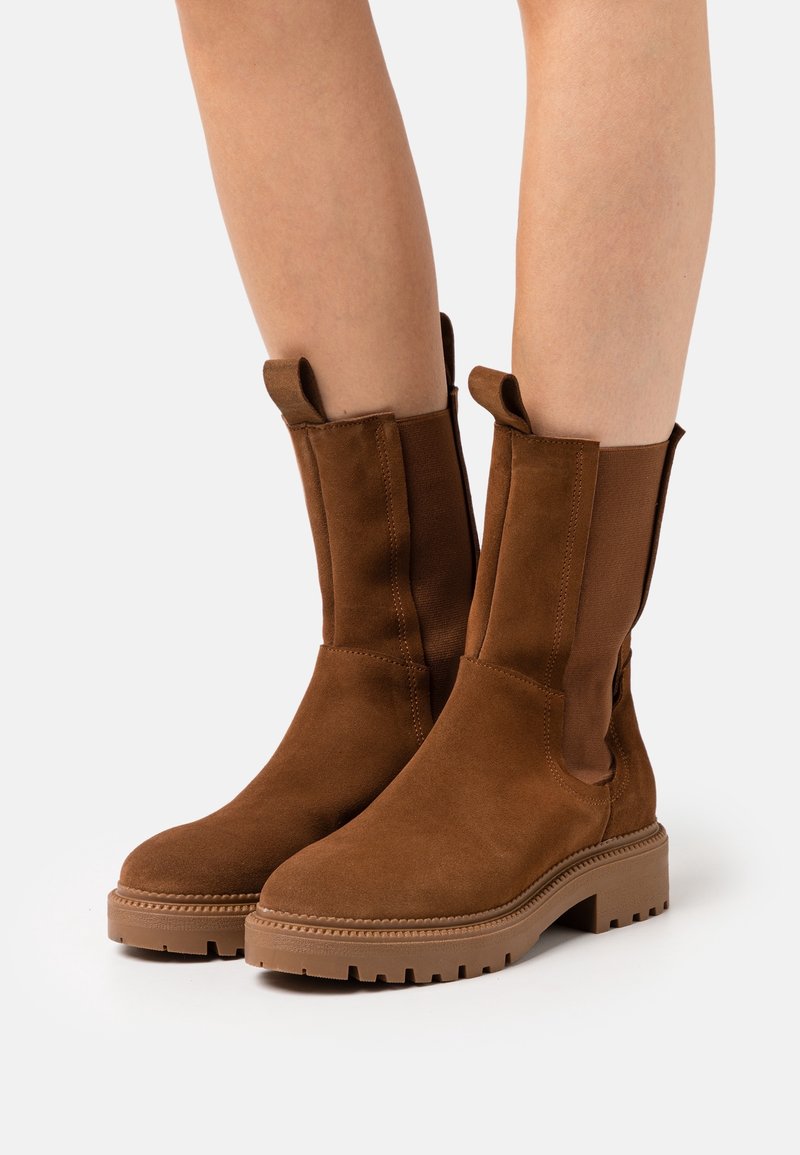 Bottes en daim marron mi-mollet avec semelles en caoutchouc épaisses portées sur des jambes nues, présentant des panneaux élastiques sur les côtés et des languettes de tirage sur un fond blanc.