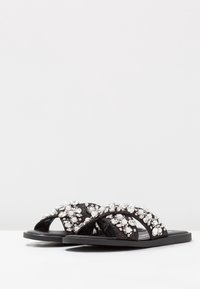 Svarta slidesandaler med en strukturerad rem prydd med klara och vita dekorativa stenar. Platt sula och öppen tå-design.