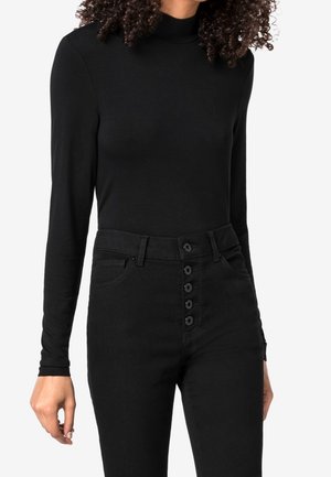 Femme portant un col roulé noir à manches longues rentré dans un jean noir taille haute avec cinq boutons visibles à l'avant.