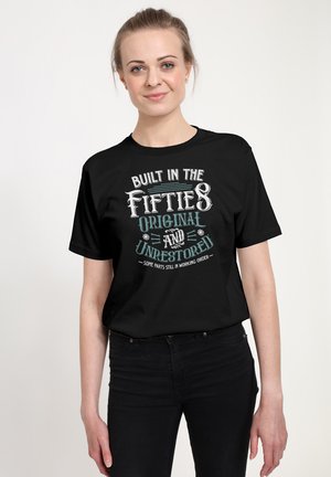 Frau mit zurückgebundenem hellen Haar, die ein schwarzes T-Shirt trägt mit der Aufschrift "Built in the fifties original and unrestored, some parts still in working order."