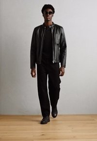 Chaqueta de bomber de cuero negro sobre una camisa negra, combinada con pantalones y zapatos negros. El modelo lleva gafas de sol oscuras y camina sobre un suelo de madera.