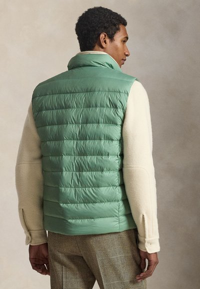 Άνδρας που φοράει ανοιχτό πράσινο καπιτονέ γιλέκο πάνω από κρεμ πουλόβερ και καρό παντελόνια, από πίσω, με ουδέτερο φόντο.