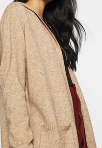 Béžový pletený kardigan s volnějším střihem, dlouhými rukávy a žebrovanými detaily na kapse. Měkká textura a jemný vzor jsou patrné.