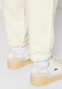 Creme sweatpants med elastiske manchetter, parret med hvide sneakers med en tan sål og grønt Lacoste-logo, fremhævet af grå ruskinddetaljer.