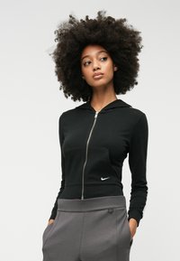 Svart zip-up hoodie med framficka, långa ärmar och en silverfärgad dragkedja. Har en liten vit Nike-logotyp på vänster sida. Grå byxor under.