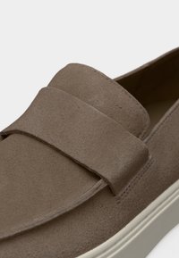 Καφέ σουέτ μοκασίνι με φαρδιά λωρίδα στο πάνω μέρος και λευκή λαστιχένια σόλα, εμφανίζεται σε κοντινή λεπτομέρεια.